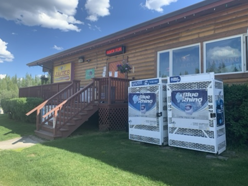 Log cabin constructed building with stairs leading to fenced in porch. Two Blue Rhino propane collec tion and dispensing cages are to the right and on the ground. The weather is warm, blue sky, puffy white clouds and perfectly trimmed green grass.